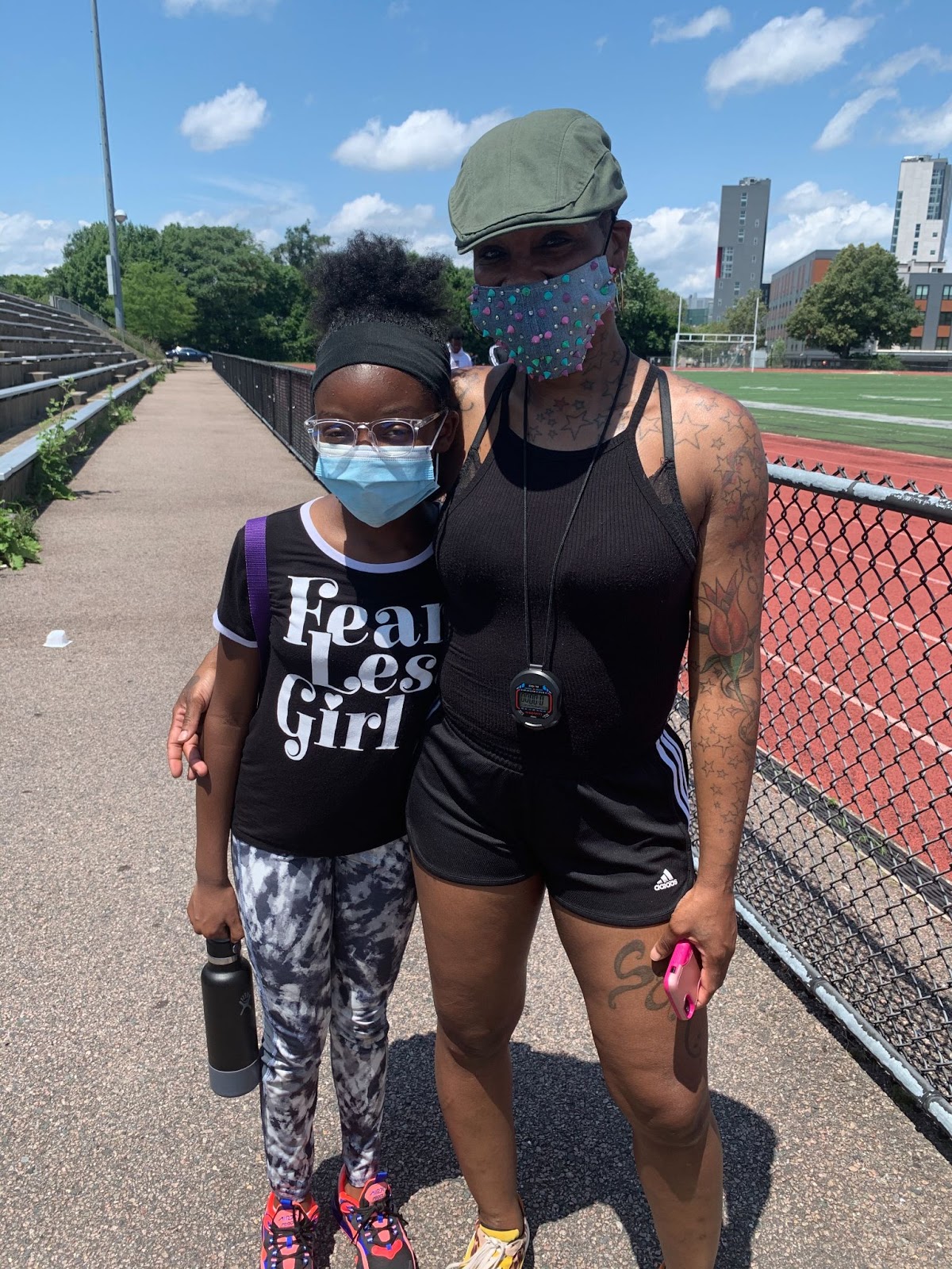 A woman standing with her arm around a little girl. An outdoor track for running is in the background, along with some trees.The sky is blue.